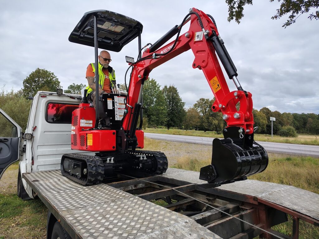 Röd minigrävare på släp med skopa och larvband, redo för transport.